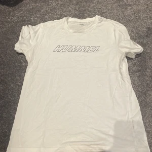 Vit Hummel t-shirt med tryck - Vit t-shirt från Hummel med klassiskt logotryck framtill och beige pilar på ryggen. Skön passform och tillverkad i mjuk bomull, perfekt för träning eller chill. Rund hals och korta ärmar gör den enkel att matcha med jeans eller shorts.