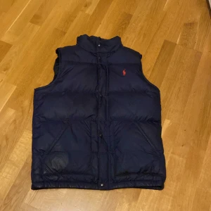 Mörkblå dunväst från Ralph Lauren - Snygg mörkblå dunväst från Ralph Lauren med klassisk röd logga på bröstet. Västen har hög krage, två snedställda fickor och stängs med både dragkedja och knappar. Perfekt för lager-på-lager och chill höst- eller vinterstil. Vesten är dock ganska gammal och har lite defekter men inget man tänker på. Luva ingår också. Hör gärna av vid intresse