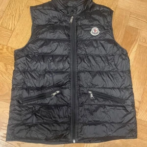 Svart dunväst från Moncler - Svart dunväst från Moncler med quiltad design och hög krage. Västen har två snedställda fickor med dragkedja och ett snyggt Moncler-märke på bröstet. Materialet är glansigt och lätt, perfekt för lager-på-lager. Stängs med dragkedja framtill. lite sönder ner till vid dragkedjan men funkar precis som vanligt 