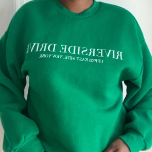 Grön sweatshirt med vit text - Säljer en grön sweatshirt med vit text på bröstet där det står 'RIVERSIDE DRIVE UPPER EAST SIDE, NEW YORK'. Tröjan har rund halsringning och långa ärmar med ribbade muddar. Perfekt för en chill och avslappnad stil.