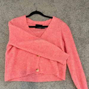 Rosa kofta från Pieces med knappar - Superfin rosa kofta från Pieces i stickat material. Koftan har v-ringning, tre stora knappar framtill och är croppad i modellen. Perfekt att slänga på sig över ett linne eller t-shirt. Långärmad och mjuk, passar både till jeans och kjol.