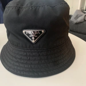 Svart bucket hat från Prada - Snygg svart bucket hat från Prada med ikonisk metalltriangel framtill. Hatten är tillverkad i återvunnen nylon och har en klassisk rund form med sömmar runt brättet. Perfekt för dig som vill ha en stilren och trendig accessoar med lyxig känsla.