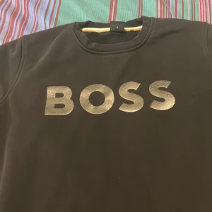 Svart sweatshirt från BOSS - Svart sweatshirt från BOSS med stor logga i silver på bröstet. Klassisk rund halsringning och ribbade muddar. Tillverkad i mjuk bomullsmix, perfekt för en clean och stilren look. Snygg detalj med liten randig etikett i sidan.