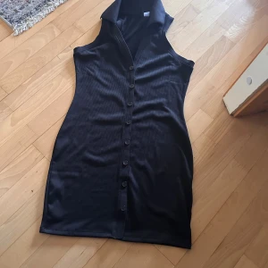 Svart ärmlös skjortklänning - Svart ärmlös skjortklänning med krage och knappar hela vägen framtill. Klänningen har en tight passform och är tillverkad i ett mjukt, ribbat material som ger en snygg siluett. Perfekt för dig som gillar minimalistisk och stilren look.