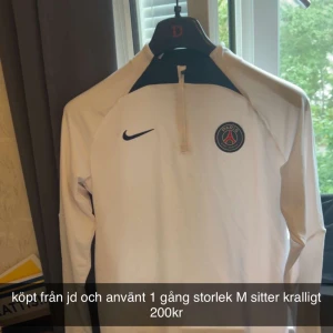 Vit långärmad PSG fotbollströja Nike - Snygg vit långärmad fotbollströja från Nike med Paris Saint-Germain-logga på bröstet och mörkblå detaljer vid axlarna. Halv dragkedja framtill och sportig passform, perfekt för träning eller match. Materialet är lätt och andas.