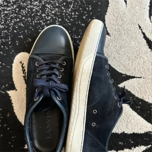Svarta sneakers från Lanvin i mocka - Stilrena svarta sneakers från Lanvin med ovandel i mocka och detaljer i skinn. Skorna har rund tå, snörning och vit platt sula. Perfekta för dig som gillar en clean och klassisk look med lyxig känsla.