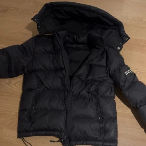 Svart pufferjacka NVLTY - Säljer en svart pufferjacka från NVLTY med huva och dragkedja. Jackan har en stilren design med långa ärmar och är perfekt för kyligare dagar. Den är både snygg och praktisk med fickor framtill. Fräsch jacka knappt använd nästan som ny.