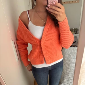 Corall kofta från Jake's, storlek S - Superfin orange/ corall kofta från Jake's i storlek S. Den är ribbstickad med v-ringning och knappar framtill. Perfekt att slänga över ett linne eller t-shirt för en chill och färgglad look. Koftan har långa ärmar och känns riktigt mjuk och skön. På bilden ser koften orage ut men i verkligheten är den mer corall färgad! 