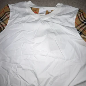 Vit Burberry t-shirt med rutiga ärmar - Snygg vit t-shirt från Burberry med klassiska rutiga detaljer på ärmarna. Rund halsringning och stilren design som ger en lyxig vibe. Perfekt för dig som gillar ikoniska märken och vill sticka ut med en clean look.