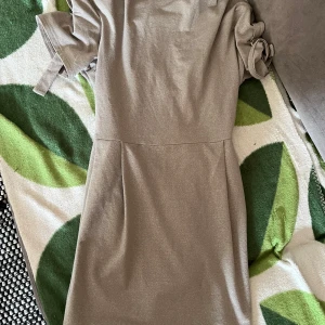 Glittrig kortklänning från Riley - Snygg glittrig kortklänning från Riley med knytband i midjan och på ärmarna. Klänningen har en enkel, rak siluett och är tillverkad i mjukt material som känns skönt mot huden. Perfekt för dig som gillar stilrena och neutrala färger.