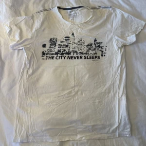 Vit t-shirt Esprit med city-tryck - Vit t-shirt från Esprit med svart tryck av en stadssilhuett och texten 'THE CITY NEVER SLEEPS' på bröstet. Klassisk rund hals och korta ärmar. Perfekt för dig som gillar urban stil och grafiska prints.