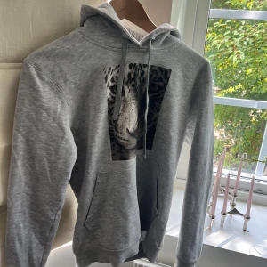 The cool elephant hoodie - Snygg grå hoodie med stor leopardbild på bröstet. Tröjan har huva med snörning och en klassisk känguruficka framtill. Perfekt för dig som gillar djurmönster och vill ha en chill och bekväm stil. Från the cool elephant 