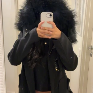 Svart parkajacka från Dennis Maglic - Svart parkajacka från Dennis Maglic med stor fluffig fuskpäls runt huvan. Jackan har dragkedja, snygga spänndetaljer och stora fickor framtill. Köpt för 1799. 