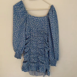 Blommig blå kortklänning med volanger - Supersöt blå kortklänning med vita små blommor, långa puffärmar och volangdetaljer framtill. Klänningen har smock vid ärmslut och en rynkad omlott-look över hela framsidan. Perfekt för dig som gillar romantisk och trendig stil.