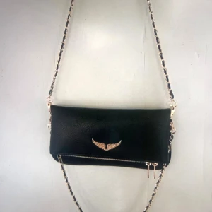 Svart axelväska från Zadig & Voltaire - Snygg svart axelväska i skinn från Zadig & Voltaire med guldfärgade detaljer, kedjerem och ikoniska vingemblemet framtill. Väskan har en praktisk dragkedja och cool struktur i materialet. Perfekt för dig som gillar stilrena accessoarer med edge.