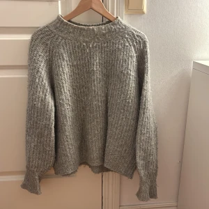 Grå stickad tröja från 157 - Mysig grå stickad tröja från 157 med grov struktur och bred ribbad krage. Tröjan har en loose passform och långa ärmar med breda muddar. Perfekt för lager-på-lager under kyliga dagar.
