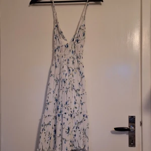 Vit blommig kortklänning med smala axelband - Supersöt vit kortklänning med blå blommigt mönster och smala axelband som korsas i ryggen. Klänningen har v-ringad front, volangdetaljer och är tillverkad i ett lätt, plisserat tyg som ger en flowy känsla. Perfekt för varma dagar och sommarhäng.