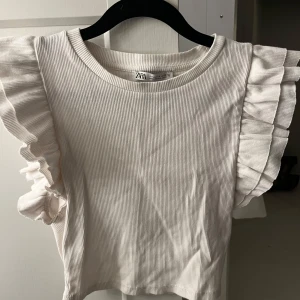 Vit ribbad topp med volangärmar Zara - Supersöt vit ribbad topp från Zara med breda volangärmar som ger en extra touch. Toppen har rund halsringning och är i ett mjukt, stretchigt material. Använd fåtal gånger