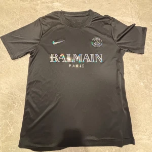 Svart PSG x Balmain fotbollströja Nike - Svart fotbollströja från Nike i samarbete med PSG och Balmain. Tröjan har korta ärmar, rund hals och är gjord i ett glansigt, sportigt material. Framsidan pryds av holografiska logotyper för Nike, Balmain och PSG. Perfekt för dig som gillar exklusiva samarbeten.