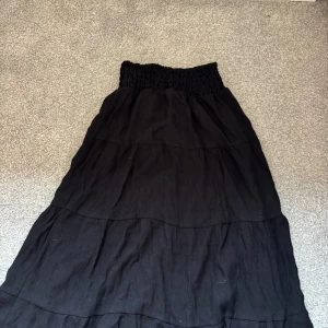 Svart långkjol från H&M med resår - Svart långkjol från H&M med bred smockresår i midjan. Kjolen har flera våder som ger en snygg och flowy känsla. Perfekt för dig som gillar en enkel men stilren look. Tillverkad i mjukt bomullstyg.