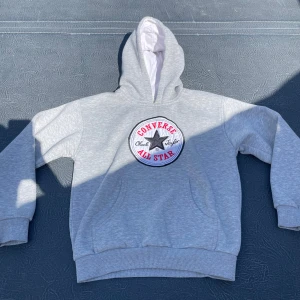Grå Converse hoodie med stor logga - Säljer en grå hoodie från Converse med stor broderad All Star-logga på bröstet. Tröjan har huva med vit insida, ribbade muddar och magficka. Tillverkad i mjuk bomullsmix (French Terry) som är skön att ha på sig. Perfekt för chill dagar och streetstyle.
