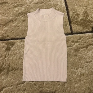Ljusbeige ribbad polotopp - Snygg ljusbeige polotopp med ribbad struktur och ärmlös design. Toppen har en hög krage som ger en cool vibe och är perfekt att styla med jeans eller kjol. Materialet är mjukt och stretchigt för en skön passform.