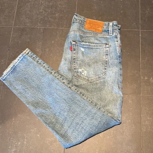 Levi's 512 ljusblå slitna jeans - Ljusblå Levi's 512 jeans med klassisk rak passform och slitna detaljer på benen för en cool, vintage look. Jeansen har fem fickor, knappgylf och den ikoniska läderpatchen bak i midjan. Perfekta för dig som gillar en avslappnad och trendig stil.