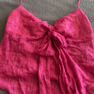 Rosa spetsig linnetopp med rosett - Supersöt rosa linnetopp med tunna axelband och stor rosett framtill. Toppen är täckt av spets och har en lätt genomskinlig look med innerfoder. Perfekt för dig som vill sticka ut med en feminin och trendig vibe. Helt ny