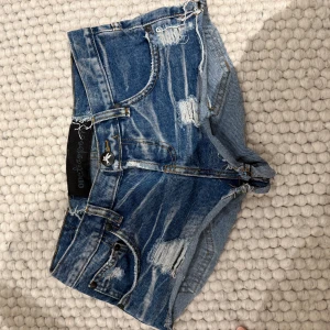 Blå slitna jeansshorts One Teaspoon - Snygga blå jeansshorts från One Teaspoon med slitna detaljer och råa kanter. Klassisk femficksmodell med knapp och dragkedja framtill. Perfekta för sommaren och ger en cool, avslappnad vibe.