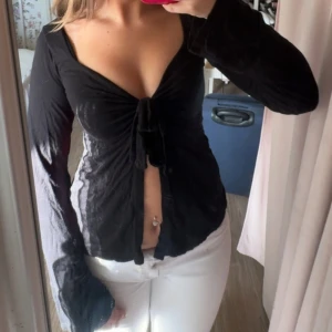 Svart knyt blus  - Supersnygg svart omlottblus från Design by Si i tunn bomull. Djup v-ringning, långa ärmar och knyt framtill som ger en feminin och trendig look. Perfekt till jeans eller kjol för en cool vibe. I storlek XS/S