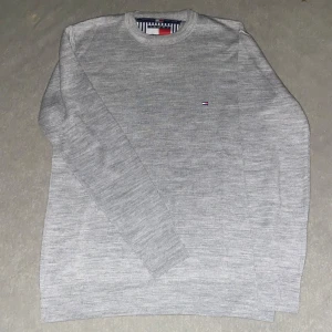 Grå stickad tröja Tommy Hilfiger - Snygg grå stickad tröja från Tommy Hilfiger med diskret broderad logga på bröstet. Klassisk rund halsringning och långärmad modell. Perfekt för lager-på-lager och enkel att matcha med jeans eller chinos.