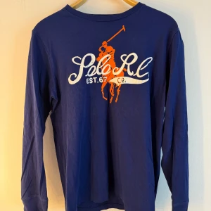 Ralph Lauren blå tröja  - Marinblå långärmad tröja från Polo Ralph Lauren med en stor orange logotyp på ryggen. Logotypen består av en polospelare till häst och texten "Polo R.L. EST. 67 CO.". Storlek L. Fint skick men lite sliten i trycket, se bilder. Något liten i storlek, mer en M
