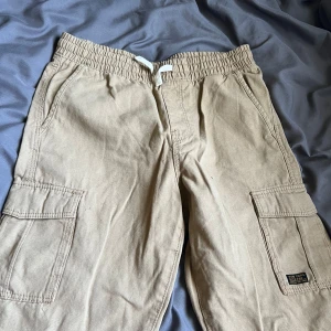 Beige cargobyxor med snörning - Säljer ett par beige cargobyxor från New Yorker med elastisk midja och vit snörning. Byxorna har stora fickor på sidorna och en bakficka med lock. Perfekta för en avslappnad och chill stil.