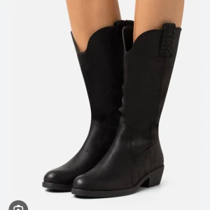 Svarta cowboy boots med låg klack - Snygga svarta cowboy boots med klassisk rund tå och låg klack. Bootsens skaft har en cool vågig kant och flätad detalj upptill. Tillverkade i skinnimitation som ger en stilren och robust känsla. Perfekta för dig som vill ha western-vibe i din outfit.