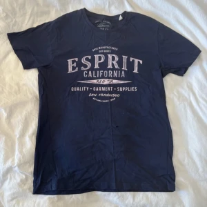 Mörkblå Esprit t-shirt med tryck - Snygg mörkblå t-shirt från Esprit med stort vintage-inspirerat tryck i beige på bröstet. Modellen är klassisk med rund hals och korta ärmar. Materialet är mjuk bomull som känns skönt mot huden. Perfekt för dig som gillar streetwear och avslappnad stil.