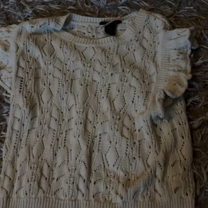 Vit stickad topp med volangärm Lindex - Supersöt vit stickad topp från Lindex med korta volangärmar och snyggt hålmönster. Perfekt för dig som gillar romantisk stil och vill ha något luftigt men ändå stickat. Rund halsringning och ribbade kanter för extra detalj.