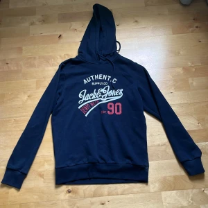 En XS - S Mörkblå hoodie från Jack & Jones - Snygg mörkblå hoodie från Jack & Jones med tryck framtill i vitt och rött. Klassisk modell med huva och dragsko, samt långa ärmar och ribbade muddar. Perfekt för chill dagar eller när du vill ha en avslappnad stil.