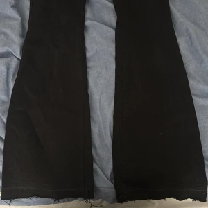 Svarta bootcut jeans från Lindex - Säljer ett par svarta jeans från Lindex i modellen Freja. De har bootcut-ben med råa, fransiga kanter längst ner och klassisk femficksdesign. Jeansen har normal passform och är tillverkade i mjuk bomull med dragkedja och knapp framtill.