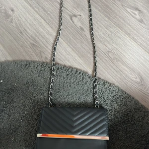 Svart quiltad axelväska med kedja - Snygg svart axelväska med quiltad ovansida och silverfärgad kedjerem. Väskan har ett stilrent metallspänne och ett insytt innerfack med dragkedja. Perfekt storlek för det viktigaste och ger en trendig vibe till din outfit.