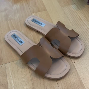 Bruna sandaler från Steve Madden - Snygga bruna sandaler från Steve Madden med breda remmar i skinn och öppen tå. Platt sula och stilren design med fyrkantig form framtill. Perfekta för varma dagar och enkel att matcha med olika outfits.
