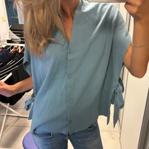 Blå skjorta med knyt i sidan - Säljer en ljusblå kofta med v-ringad hals och knappar framtill samt rosetter på sidan 