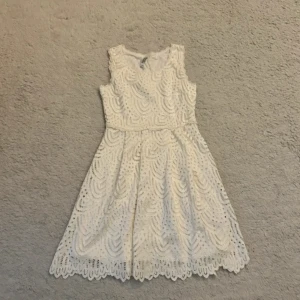 Vit spetsklänning från Chelsea Peretti - Supersöt vit kortklänning från Chelsea Peretti med spetsdetaljer och ett unikt mönster. Klänningen är ärmlös och har en rund halsringning. Perfekt för sommarens alla tillfällen! Spetsen har fina vågiga kanter och ett snyggt broderat mönster.