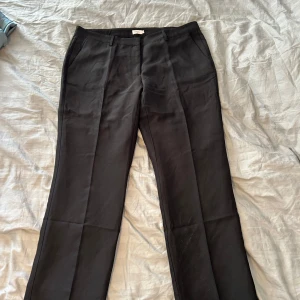 Svarta kostymbyxor från Nelly - Klassiska svarta kostymbyxor från Nelly med raka ben och pressveck. Byxorna har hällor för bälte och sidfickor, vilket ger en clean och stilren look. Perfekta för dig som gillar en enkel men snygg stil.