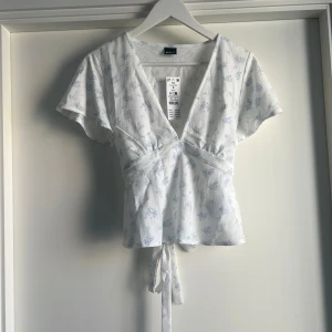 Blommig omlottopp från Gina Tricot - Supersöt vit omlottopp från Gina Tricot med ljusblått blommönster. Djup v-ringning, korta ärmar och knytband i midjan för en snygg siluett. Perfekt för dig som gillar romantisk och fräsch stil. Tillverkad i mjuk bomull. 280kr nypris slutsåld online. 