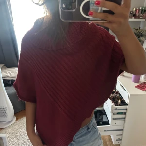 Vinröd stickad poncho med krage - Säljer en vinröd stickad poncho med bred krage och ribbad struktur. Perfekt att slänga över en t-shirt för en chill och snygg look. Asymmetrisk form och korta ärmar ger en cool vibe. Passar dig som gillar att sticka ut lite extra.