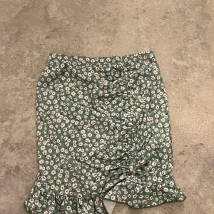Grön blommig volangkjol från Shein - Supersöt grön kjol från Shein med vitt blommönster och små röda detaljer. Kjolen har volangkant nedtill och snygg rynkning på ena sidan som ger en cool look. Perfekt för dig som gillar färg och mönster!