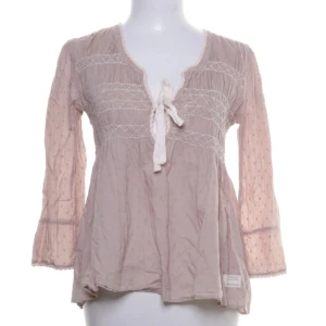 Odd Molly blus  - Supersöt rosa blus från Odd Molly med broderade detaljer och knyt framtill. Blusen har v-ringning, lätt utsvängda ärmar och ett luftigt, romantiskt fall. Perfekt för dig som gillar boho-vibe och mjuka material.