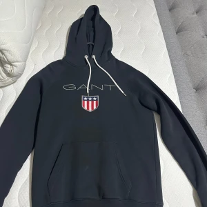 Svart hoodie från GANT med logga - Svart hoodie från GANT med vit snörning i huvan och stor broderad GANT-logga samt sköld på bröstet. Klassisk känguruficka framtill och långa ärmar. Perfekt för en chill och stilren look.