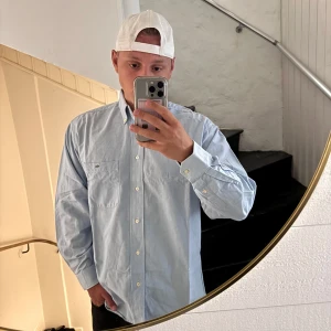 Ljusblå skjorta från Lacoste - Snygg ljusblå skjorta från Lacoste med klassisk krage och vita knappar framtill. Skjortan har en bröstficka med den ikoniska krokodilloggan och är långärmad. Perfekt för dig som gillar stilrena och tidlösa plagg.