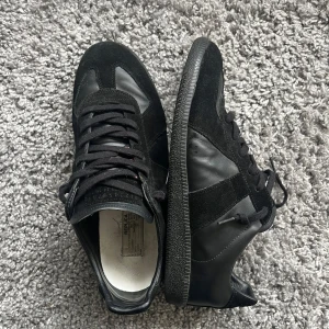 Svarta Maison Margiela  Gats🐈‍⬛ - Säljer ett par riktigt feta Maison Margiela Gats i storlek 42 (passar 43). Skorna är i bra skick, dem har blivit lite creasade samt att dem har en blekt fläck på ena skosnöret. Går att vända skosnöret åt andra håller för att de ej ska synas eller köpa nya för en liten slant. Har digitalt kvitto på skorna! Tveka ej på att höra av dig vid funderingar, inga frågor är dumma frågor! Tack!🙌🏼🤩